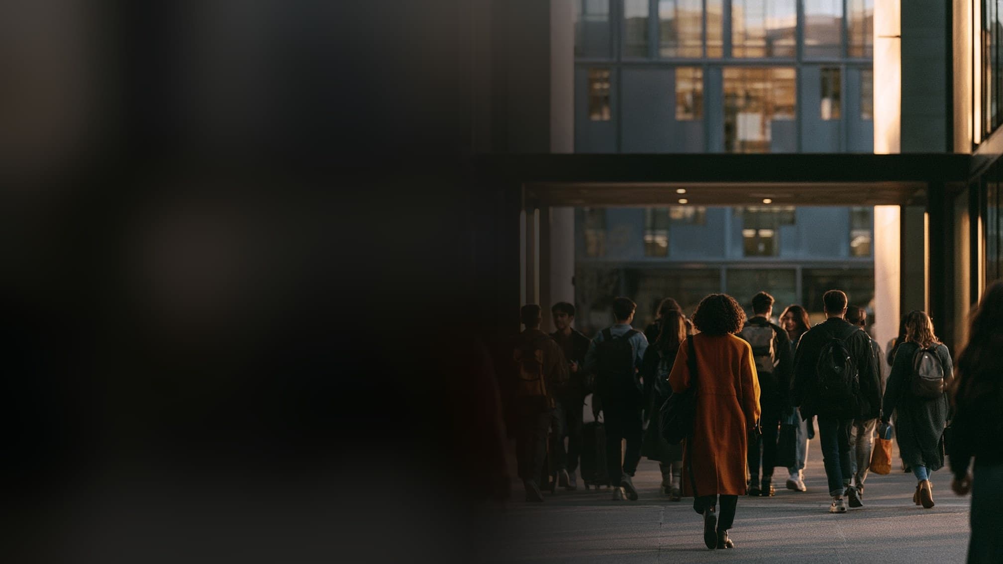 A student in a warm coat walks toward a modern university building at dusk